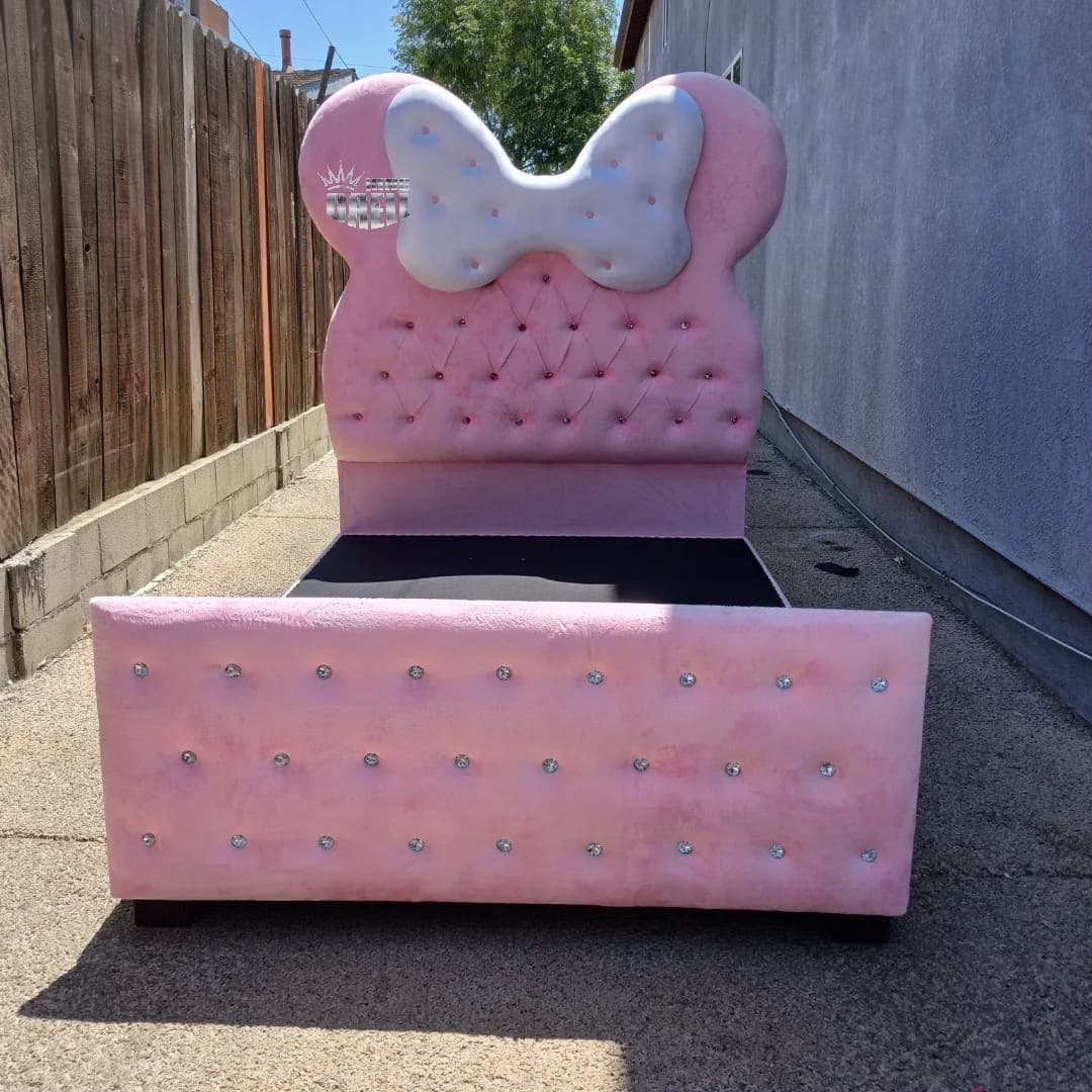 Minnie Mouse Bed head-on, its pink plush headboard topped with round ears and a white sculpted bow above diamond tufting set with crystal-style buttons